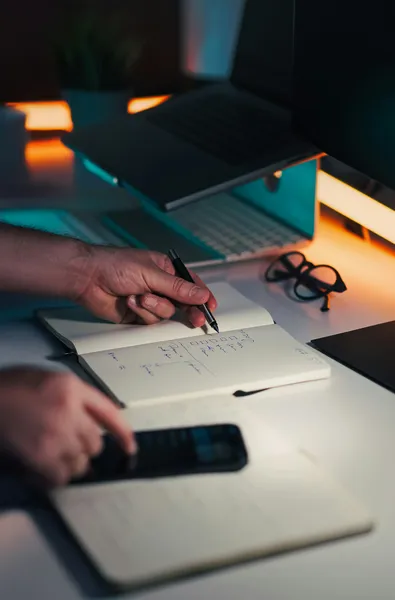 A person's hand holding a pen over an open notebook with a to-do list, beside a laptop, glasses and a phone on a desk