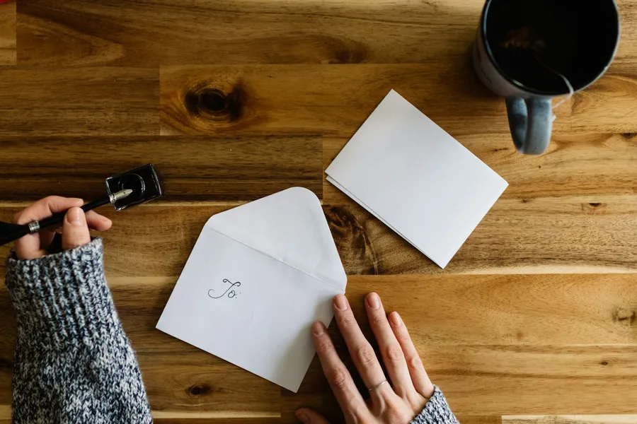 Overhead view of hands holding a pen over an envelope marked To on a wooden desk, with a mug of tea nearby