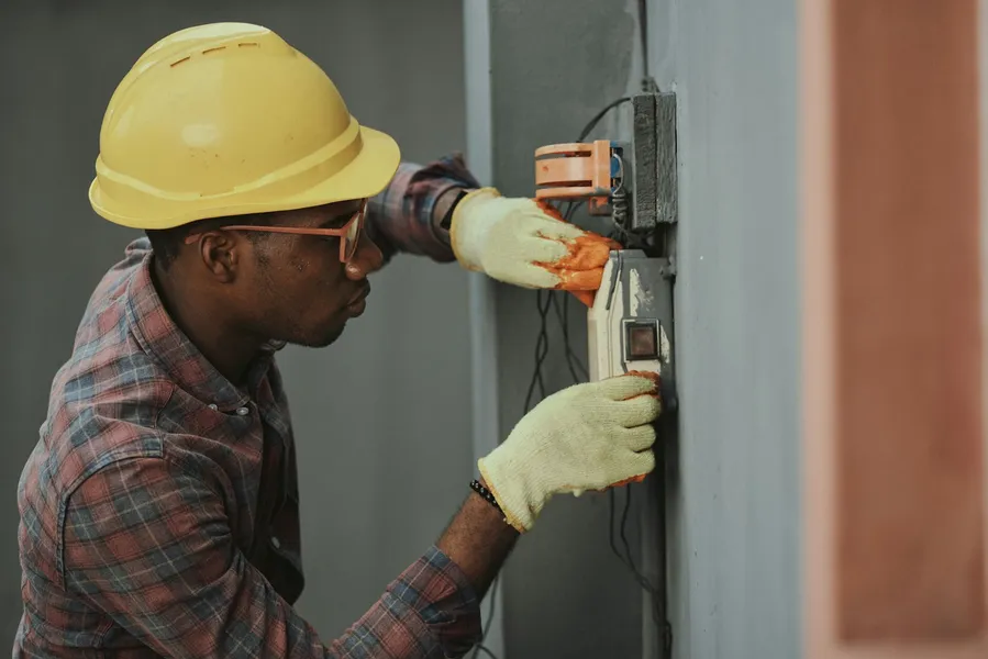 An electrician in a yellow hard hat and work gloves wiring a wall-mounted switch box