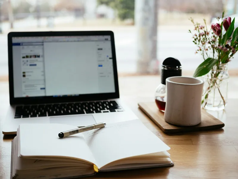 Open laptop beside a notebook, pen and mug on a wooden table by a window