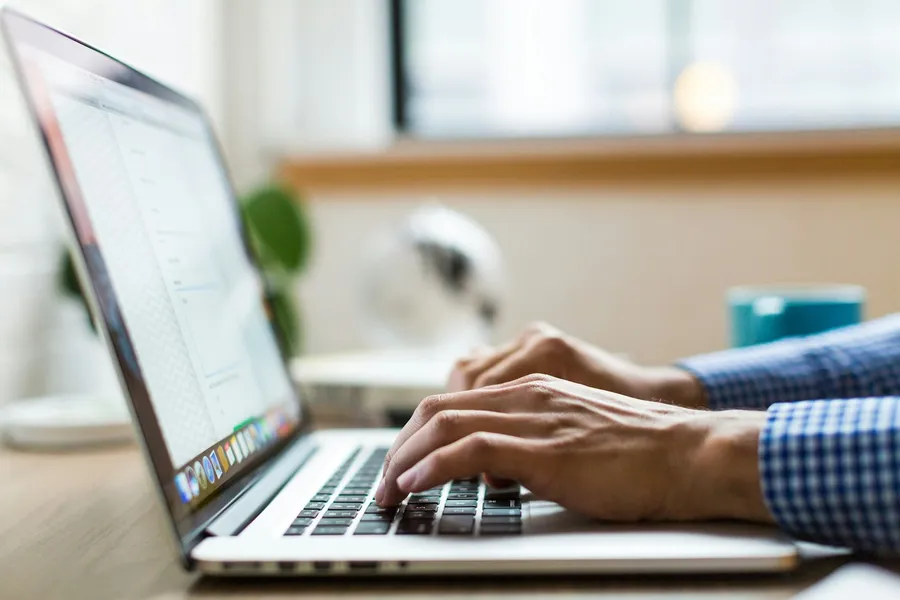 Close-up of a person's hands typing on a silver laptop at a desk by a window